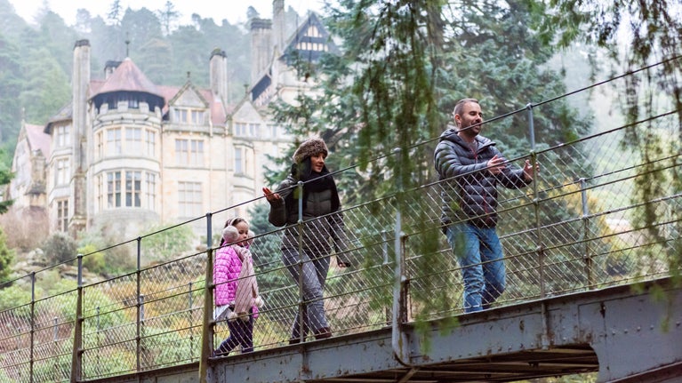 A couple with their young child walk across an iron bridge, with the large house at Cragside visible behind them in the background surrounded by evergreen trees.